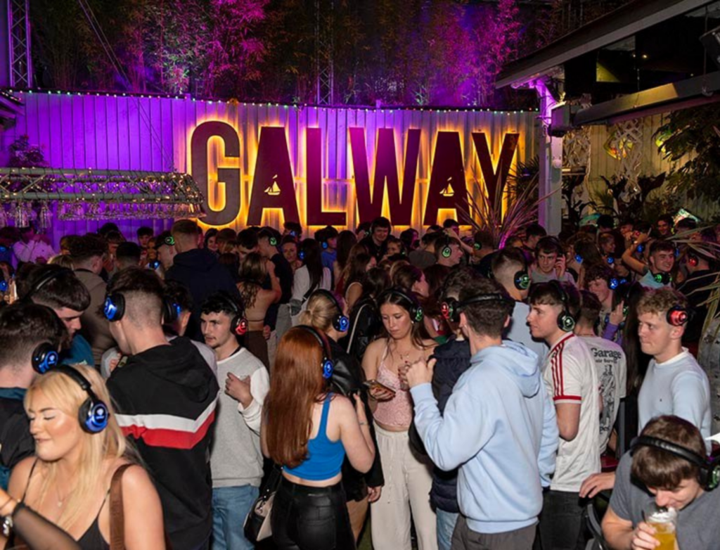 A lively silent disco event at An Púcán’s outdoor area, with a large illuminated "GALWAY" sign in the background and vibrant purple lighting setting the scene. The crowd is wearing glowing headphones in different colors, dancing and socialising in the energetic atmosphere. The space is surrounded by greenery and decorative lighting, creating a vibrant and festive vibe.