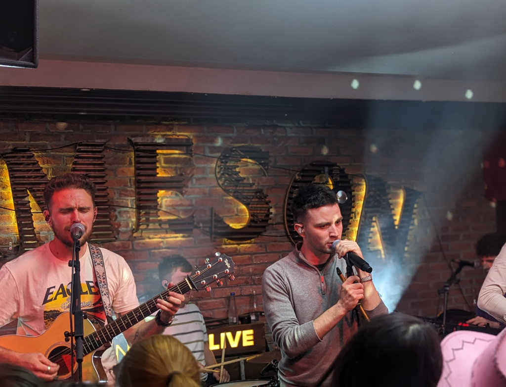 Two musicians performing on a stage with a brick wall background featuring the illuminated "JAMESON" sign. The man on the left is singing while playing an acoustic guitar, and the man on the right is holding a microphone and singing passionately. Stage lighting and a small crowd of onlookers are visible in the foreground, creating a lively atmosphere.