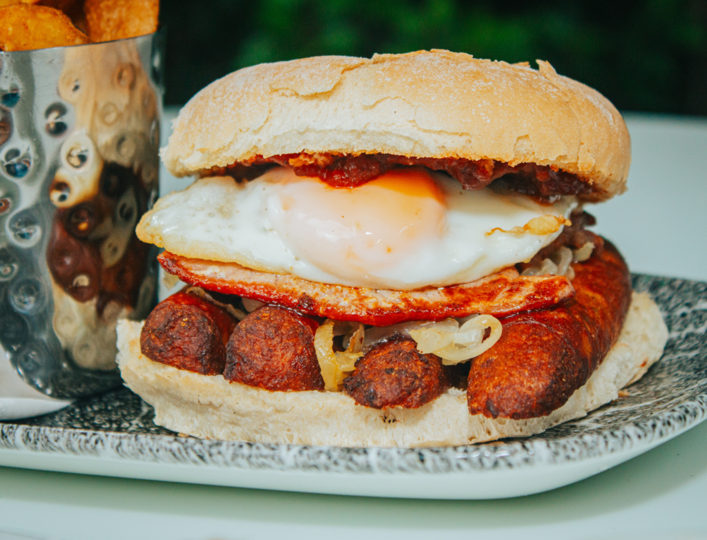 A hearty breakfast sandwich served on a floury bap, featuring crispy sausages, fried onions, a perfectly cooked sunny-side-up egg, and a dollop of tomato sauce. The sandwich is plated on a speckled dish alongside a metal cup filled with golden, chunky chips.