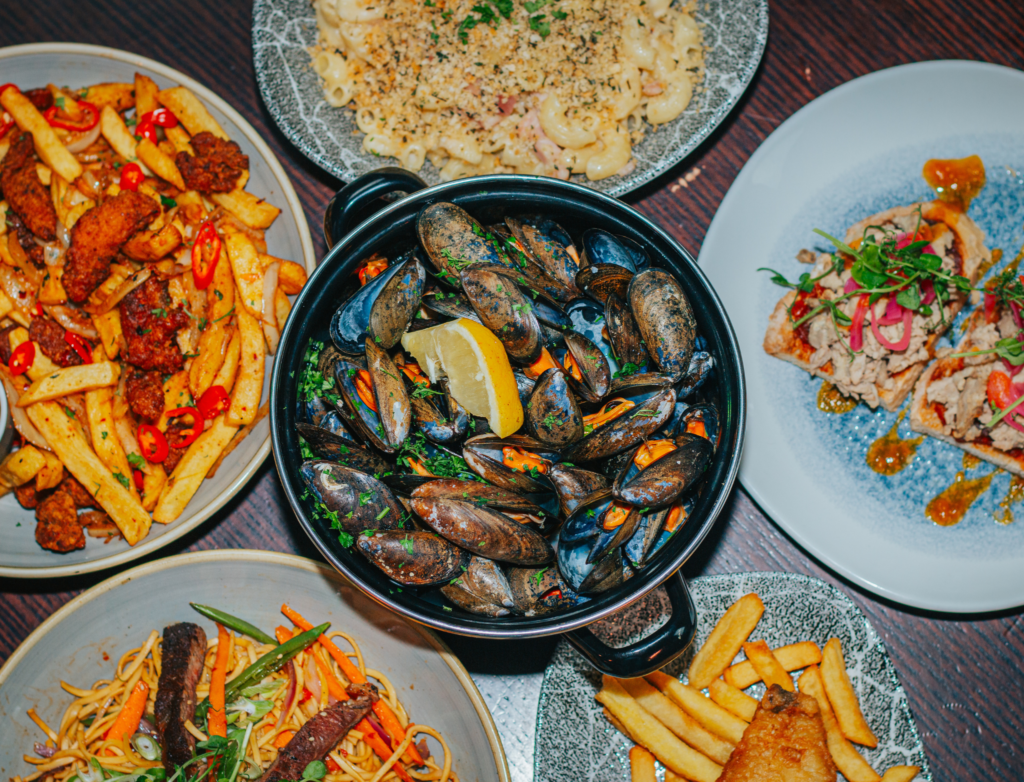 A top-down view of a table filled with a variety of dishes, including a black pot of mussels garnished with lemon wedges in the center. Surrounding it are plates of crispy fries topped with fried chicken and chili slices, mac and cheese, a seafood dish with sauce and microgreens, a noodle dish with vegetables and beef, and fish and chips. The vibrant colors and diverse textures create an inviting dining scene.