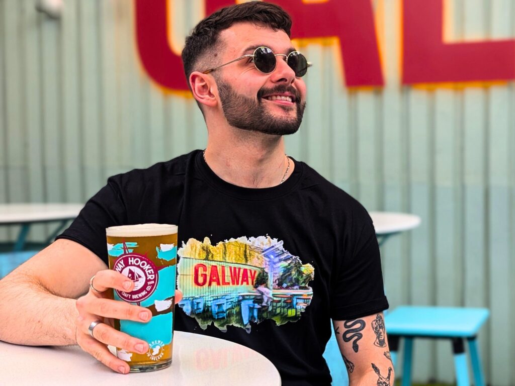 photograph of male model with a pint of galway hooker in hand showcasing branded merchandise in the form of a black t-shirt with artwork of an pucan's beer garden on the front. the model is sitting in the beer garden of an pucan