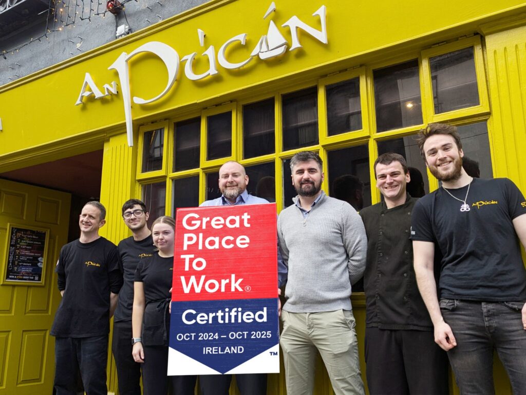 The team of An Púcán proudly standing in front of the bar's bright yellow facade with the An Púcán logo above. They are holding a "Great Place to Work Certified" sign for October 2024 to October 2025, celebrating their achievement. The group is smiling, showcasing their friendly and welcoming spirit, with the cosy and iconic entrance of An Púcán as the backdrop.