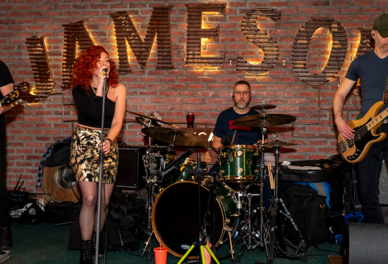 A lively band performing in front of a brick wall adorned with the illuminated "Jameson" logo. The female vocalist with vibrant red hair stands at the microphone, passionately singing, while the drummer and bassist play their instruments in the background. The setting exudes energy and excitement, perfect for a night of live music at the pub.