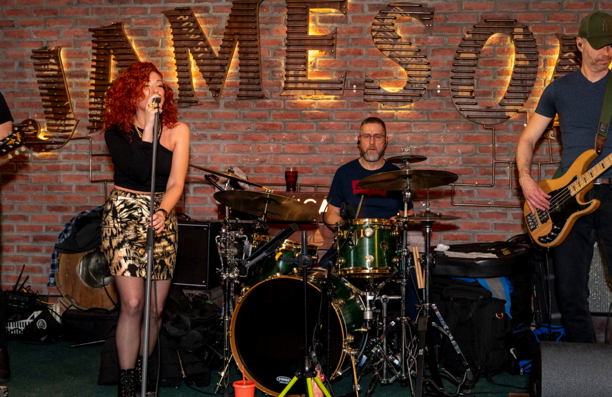 52 A live band performing at An Púcán, set against a brick wall with a glowing "JAMESON" sign. The singer, with red curly hair, stands at the microphone, while the drummer and bassist play their instruments. The scene captures a lively and intimate musical performance, with professional equipment and a cosy pub ambiance.