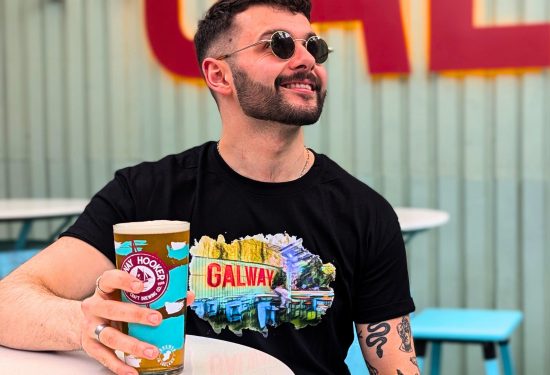 A man sitting outdoors at An Púcán, wearing sunglasses and a black t-shirt with a colorful Galway design. He is holding a pint of Galway Hooker beer and smiling as he looks off into the distance. The background features light-colored walls with bold red lettering and blue outdoor furniture, creating a relaxed and vibrant atmosphere.