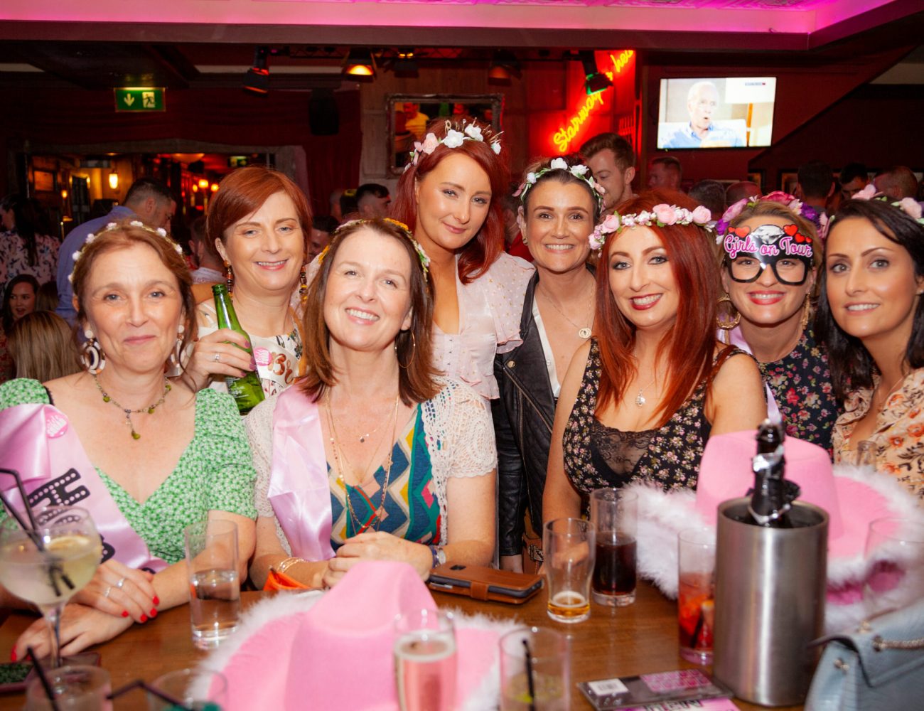 A group of women enjoying a celebration at An Púcán pub. They are seated around a table adorned with drinks and pink hen party props, including hats and sashes. Some of the women wear flower crowns, while one has novelty glasses that read "Bride to Be." Everyone is smiling and having fun in the lively pub atmosphere, with other patrons and neon signs visible in the background.