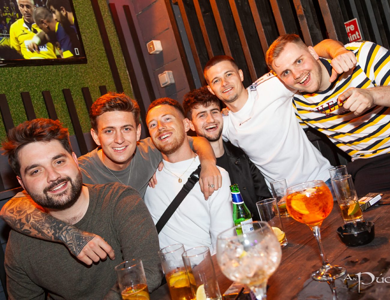A group of six friends enjoying a night out at An Púcán pub. They are seated around a table with drinks, including beer, cocktails, and mixers, smiling and laughing for the camera. One of the men has his arm around his friend, and another points toward the camera. In the background, a TV screen displays a football match, adding to the lively atmosphere. The setting features modern décor with wooden paneling and greenery.
